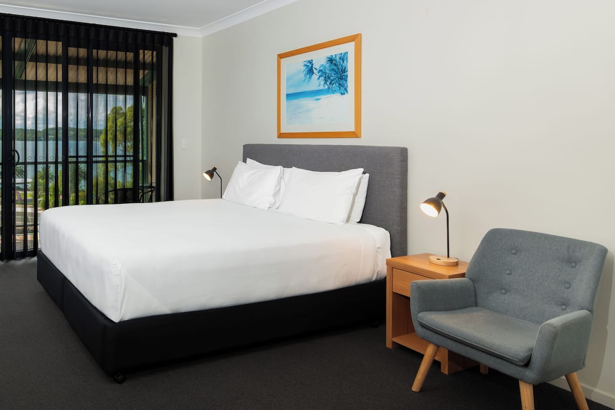 A hotel room with a large bed, a grey armchair, a nightstand with a lamp, and a framed picture on the wall.