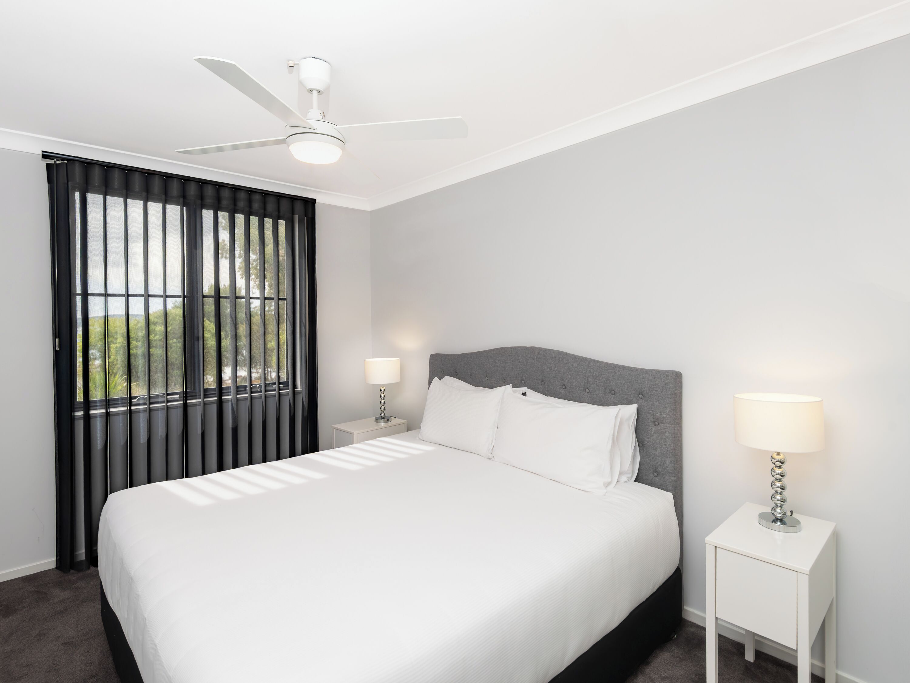 Un dormitorio moderno con una cama, dos mesitas de noche, un ventilador de techo y una ventana con cortinas.