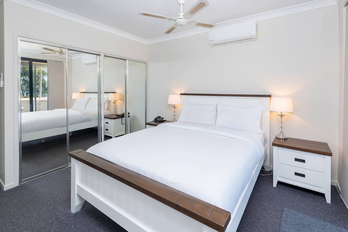 A bedroom with a large bed, a ceiling fan, a bedside table with a lamp, and a mirror.