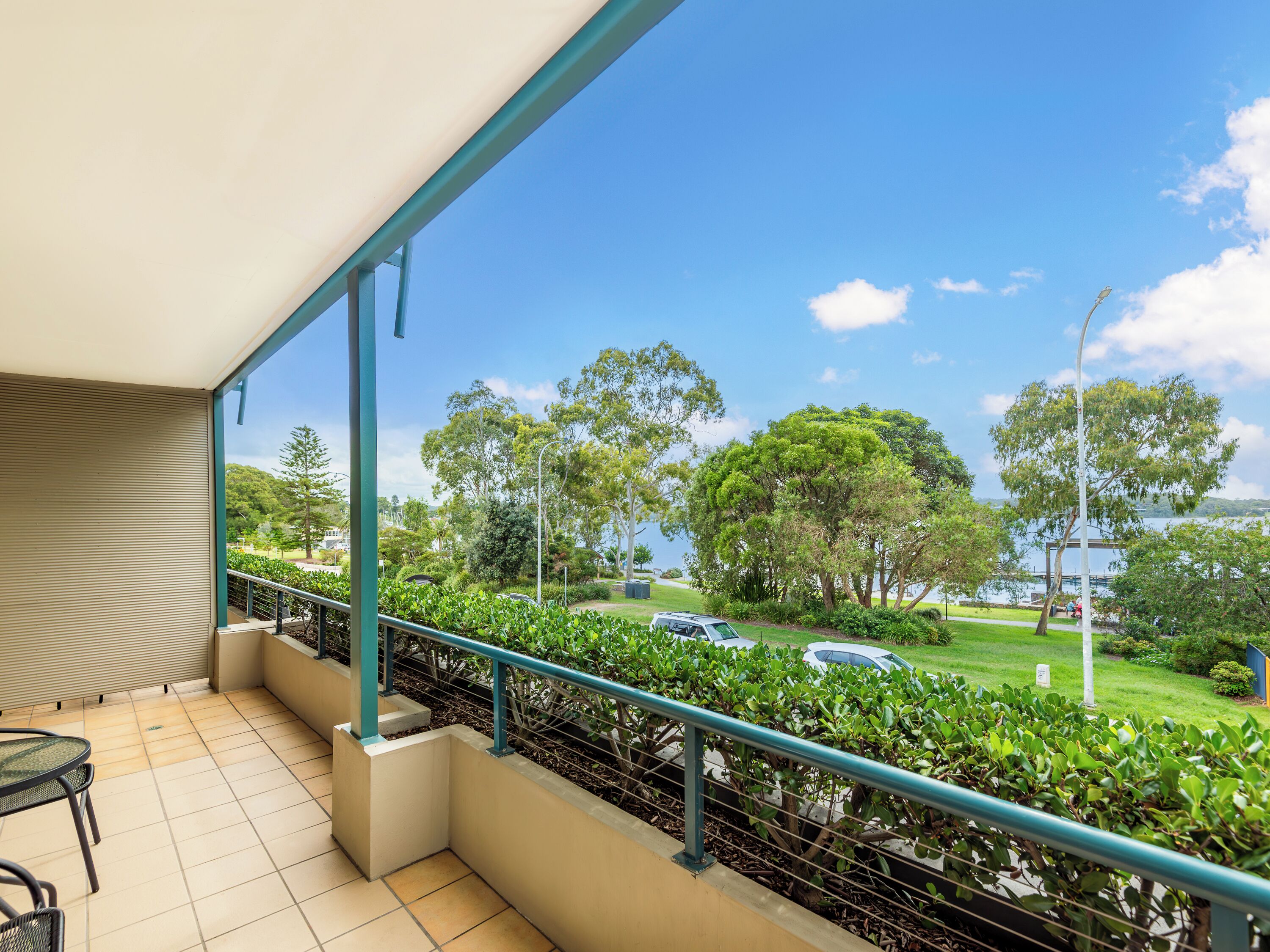 A balcony with a view of a park, a car, and a building across the street.