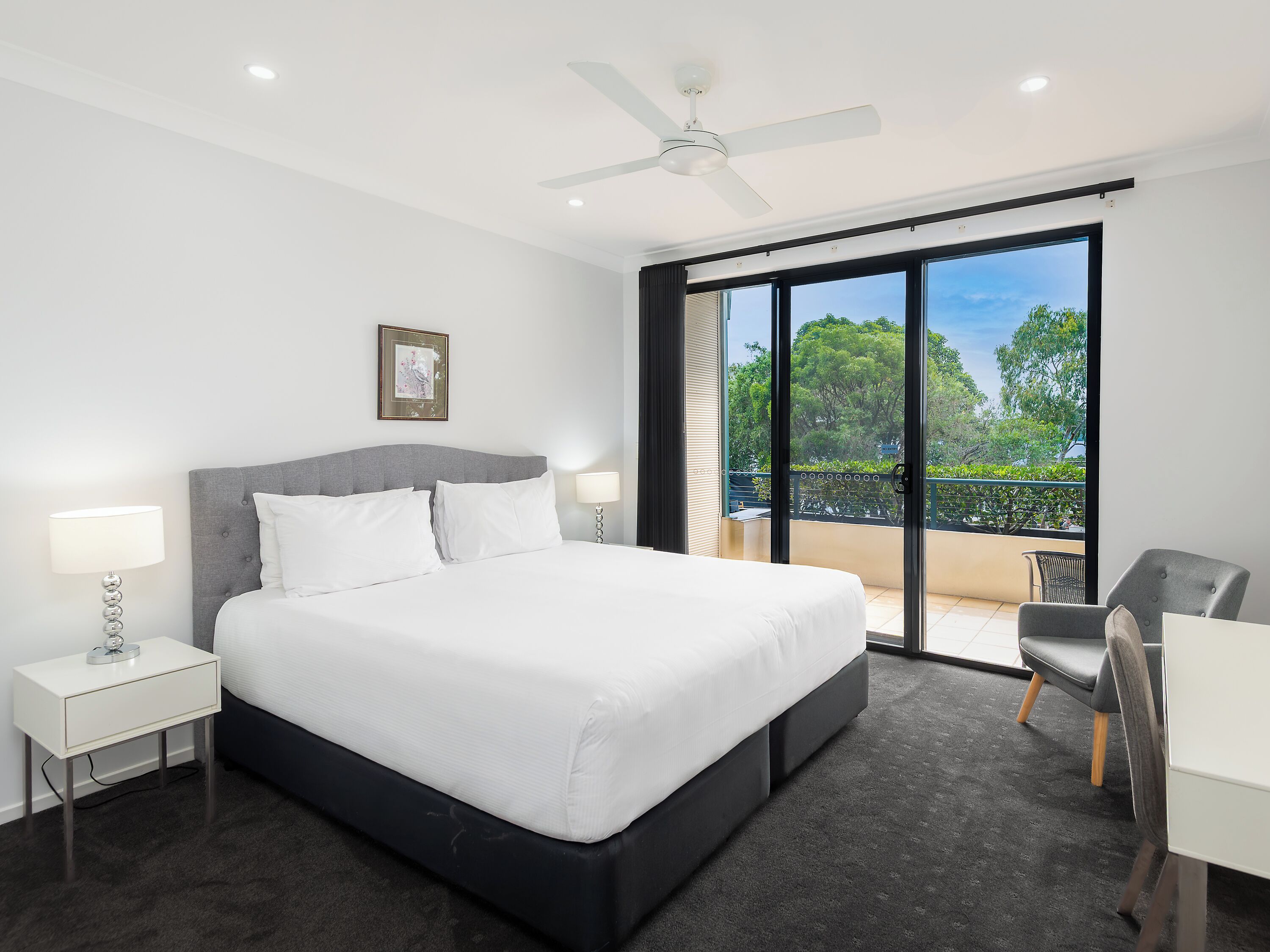 A modern hotel room with a large bed, a ceiling fan, a view of the outdoors, and a small desk with a chair.