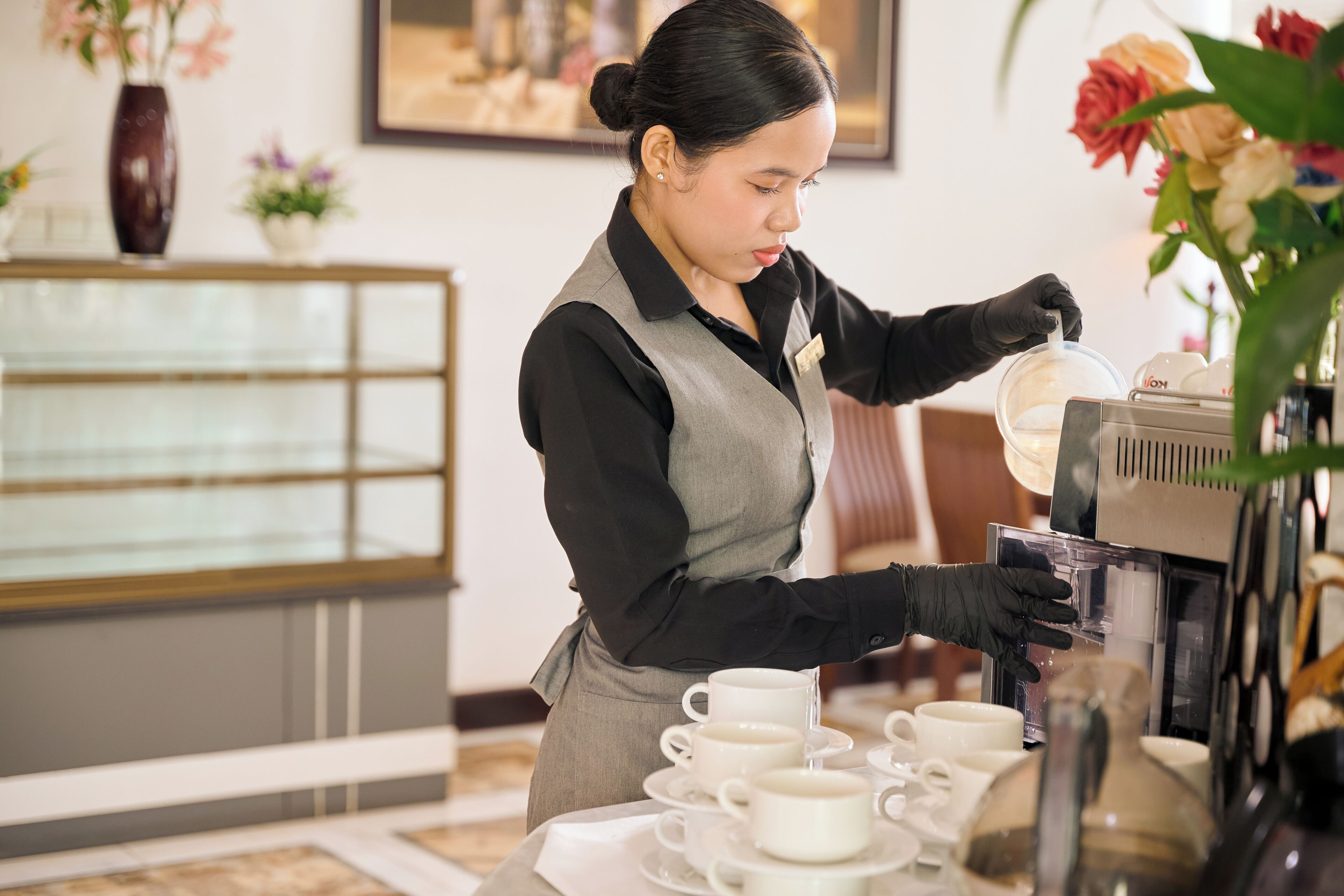 Une personne préparant du café dans un cadre professionnel, à l’aide d’une machine à café, de tasses et d’un vase décoratif.
