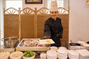 A chef in a professional kitchen with a buffet setup, including various food items and cooking utensils.