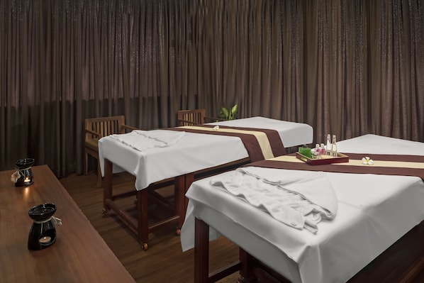 A room with two massage tables, each with a white sheet and a brown headrest. There are two candles on a wooden table, and a small plant on one of the massage tables.