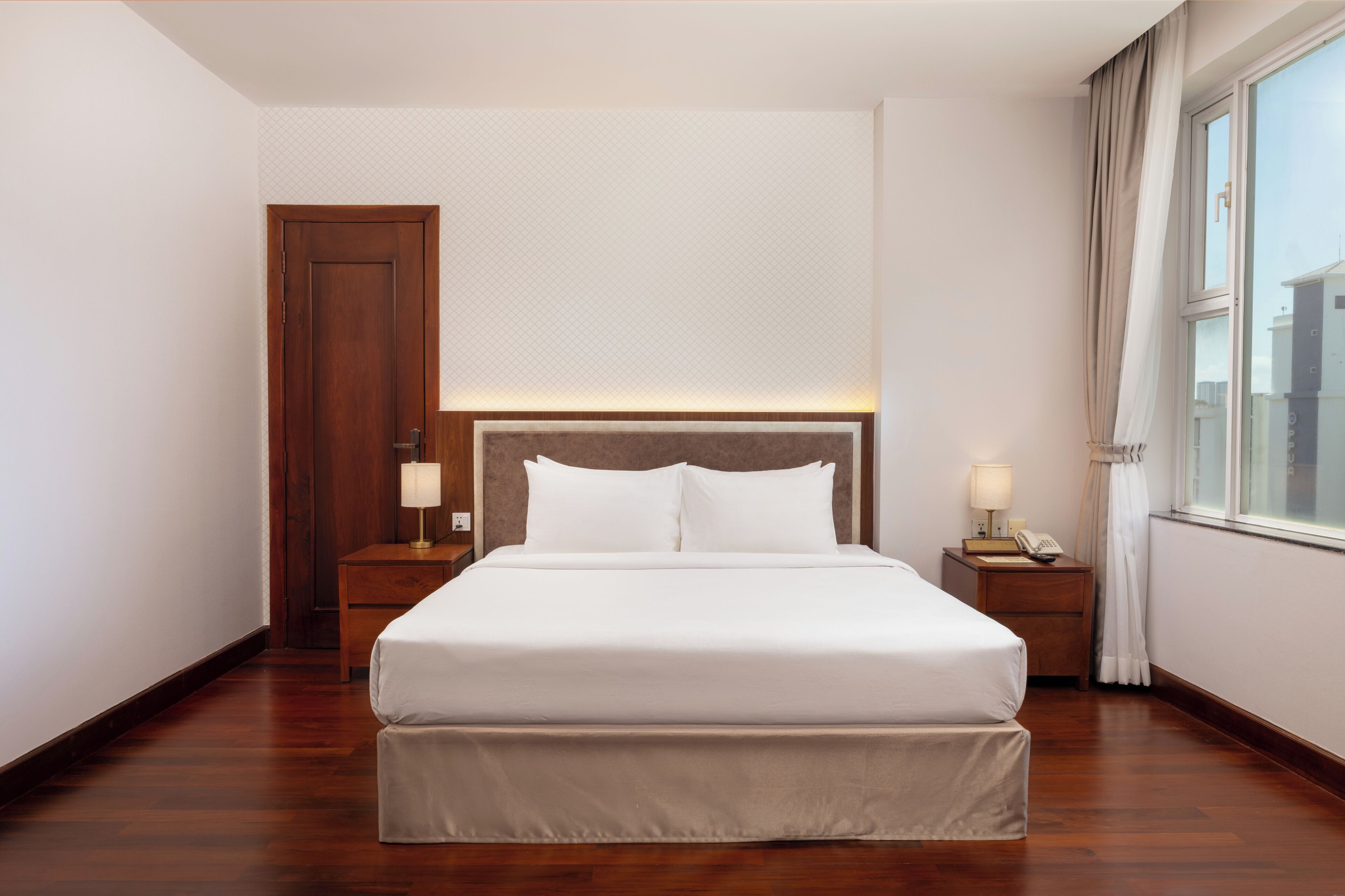 A hotel room with a large bed, wooden headboard, bedside tables with lamps, and a window with curtains.