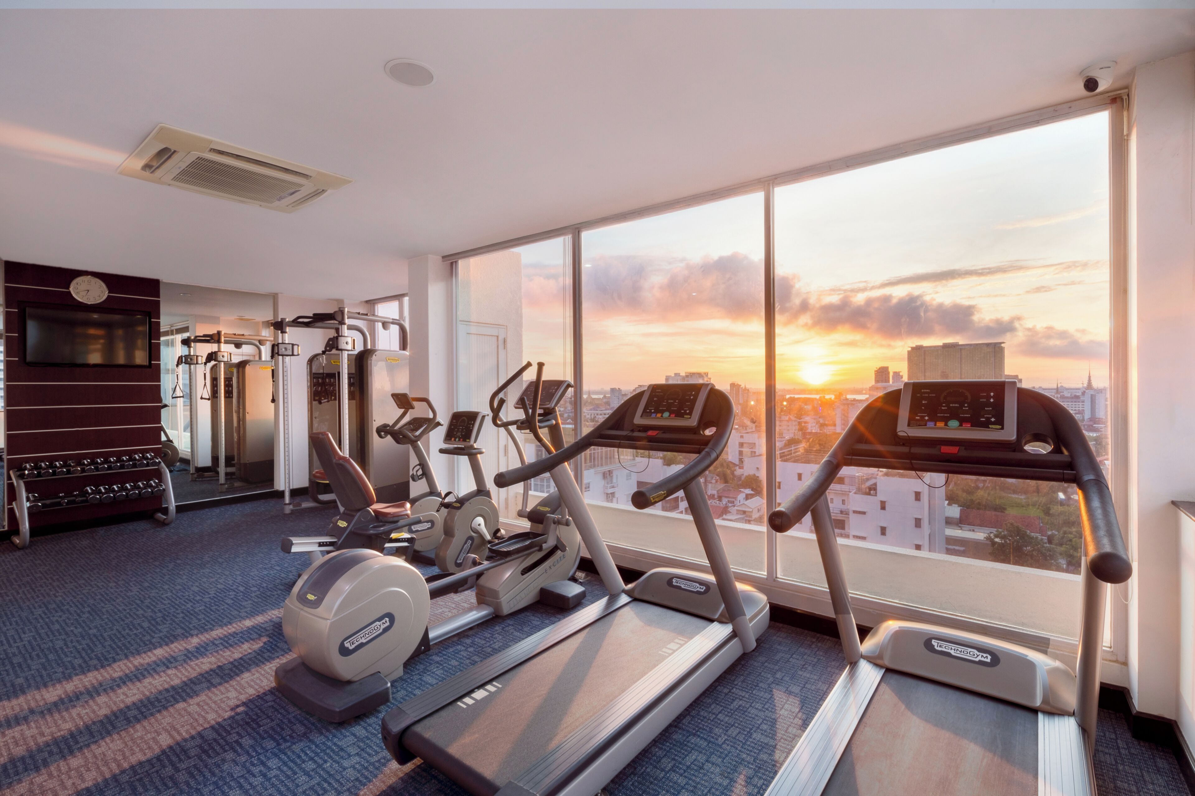 Un gimnasio con cintas de correr, elípticas y equipo de pesas, con vistas a la ciudad a través de amplios ventanales.