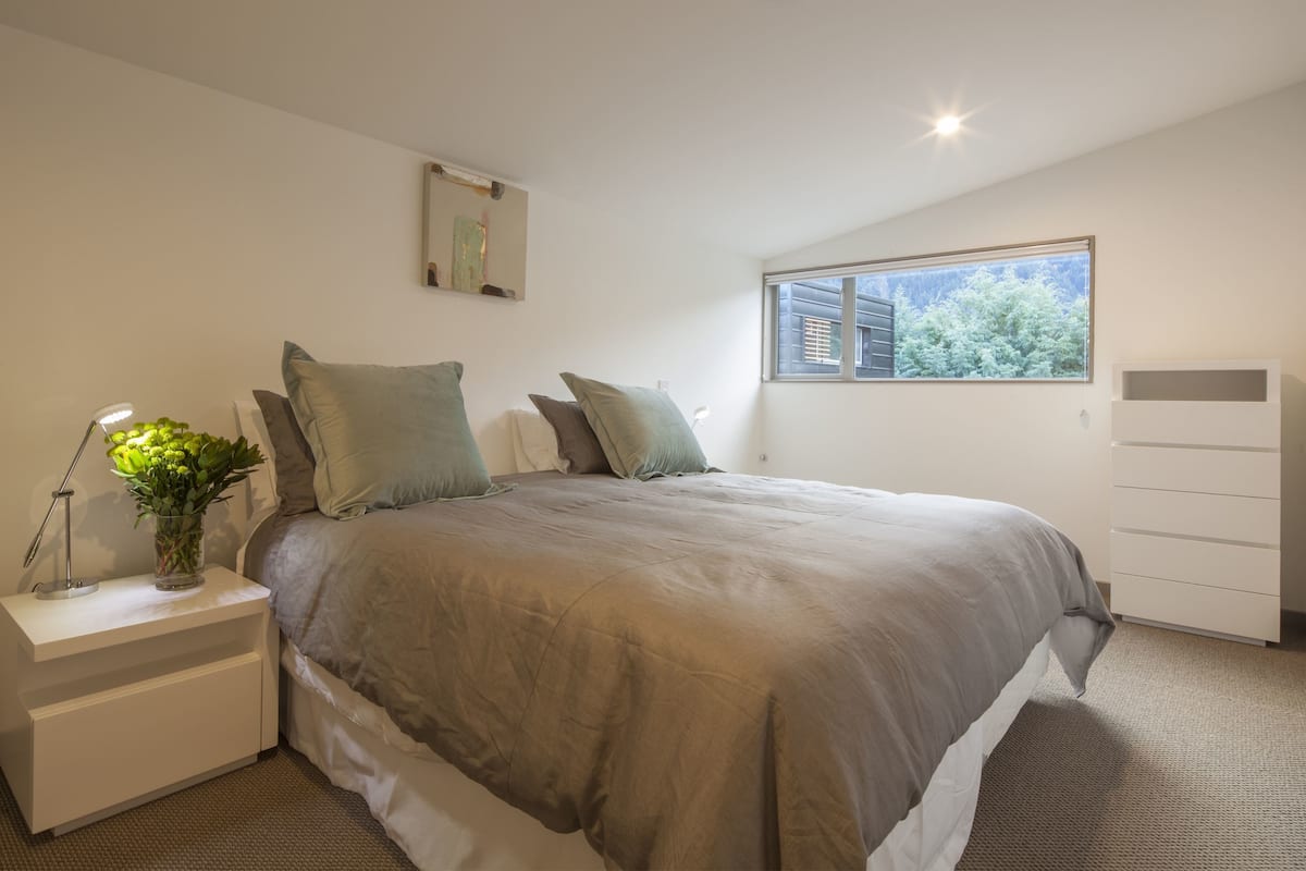 A bedroom with a large bed, a nightstand with a lamp and a vase of flowers, a dresser, and a window with a view of trees.