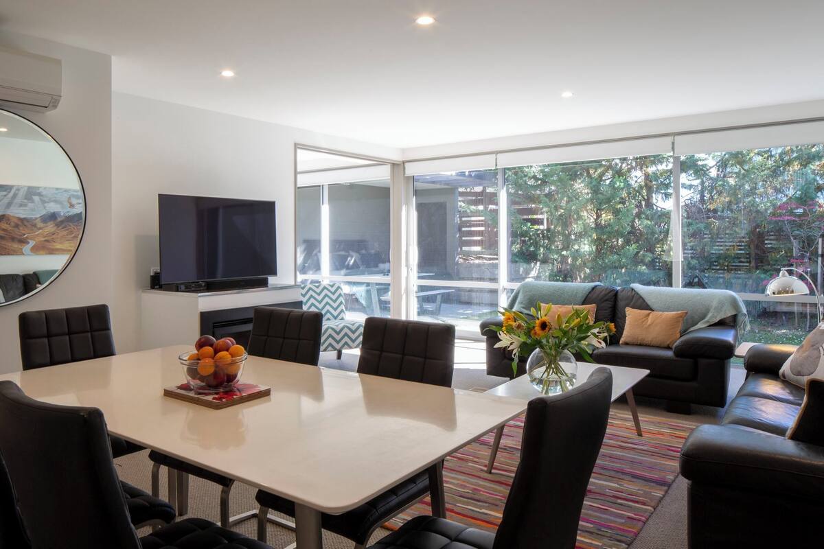 A modern living room with a dining table, chairs, a flat-screen TV, and a large window offering a view of greenery and buildings.