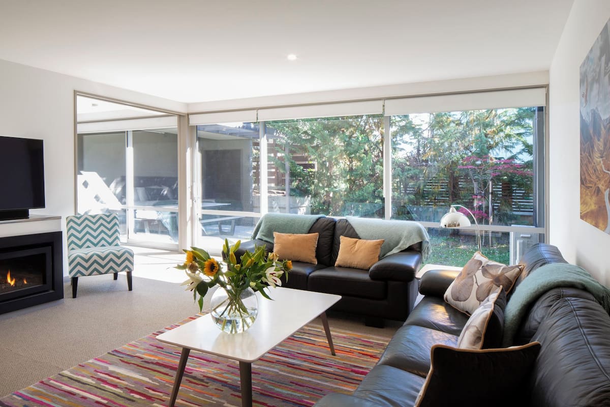 A modern living room with a fireplace, a sofa, a coffee table, and a large window offering a view of the outdoors.