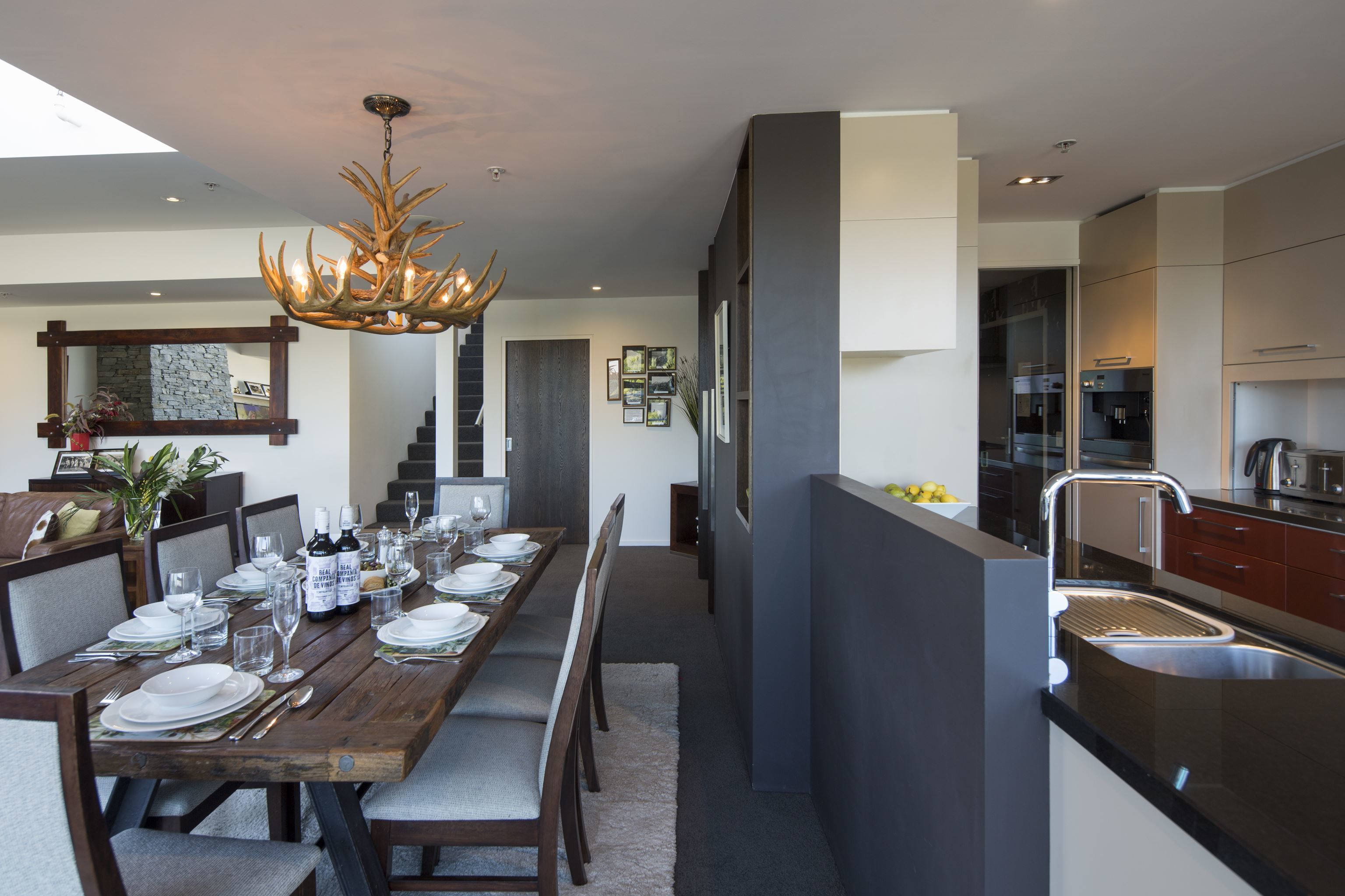 A modern kitchen with a dining area, featuring a wooden table set for six, a chandelier, and a built-in island with a sink.