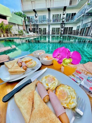 A breakfast setting with toast, eggs, and ham on a wooden table by a poolside, with a view of a modern building and a clear blue pool. - Anantra Resort Pattaya (Pattaya)