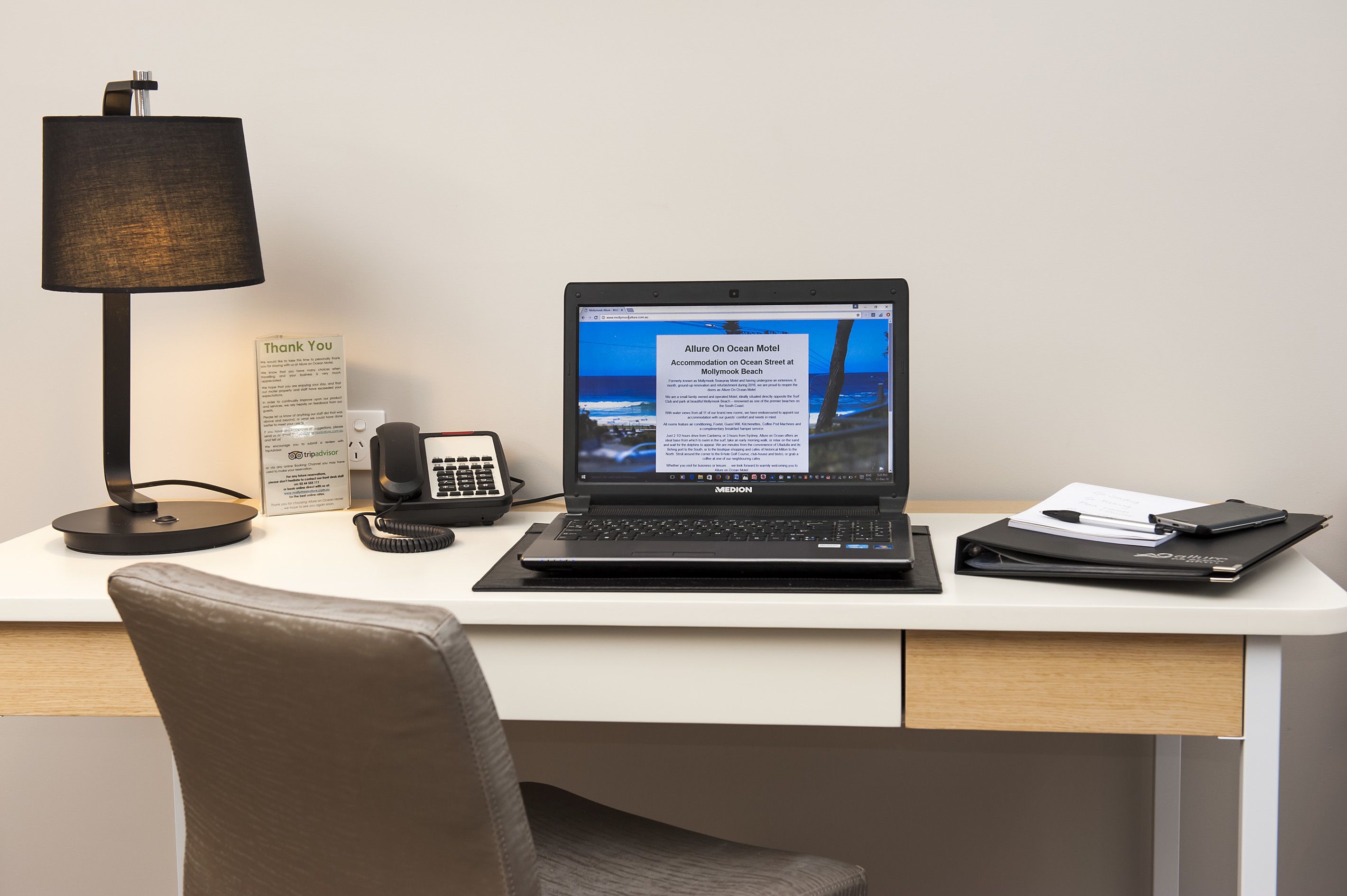 Un escritorio de hotel con una computadora portátil, un teléfono, una lámpara y un cartel.