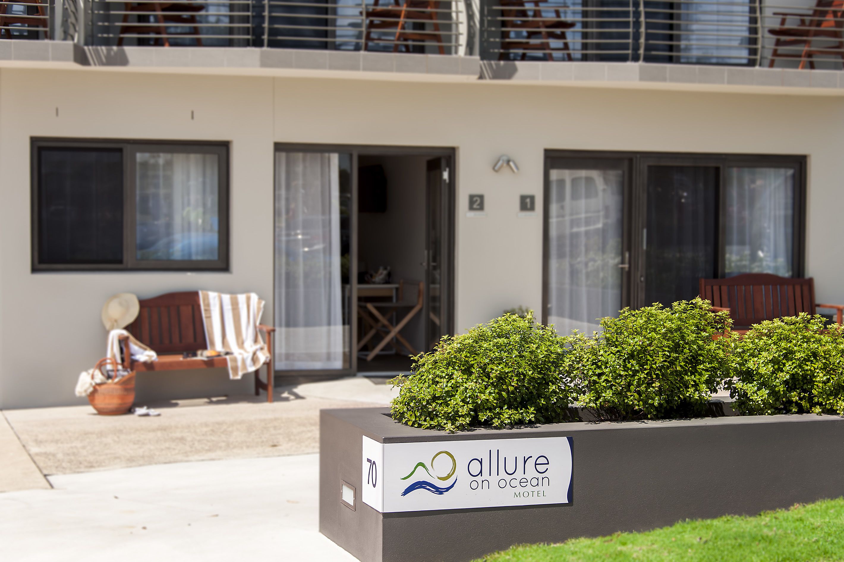 A modern building with a sign for 'Allure on Ocean Motel'. There is a bench with a hat and a basket, and a planter with green shrubs in front.
