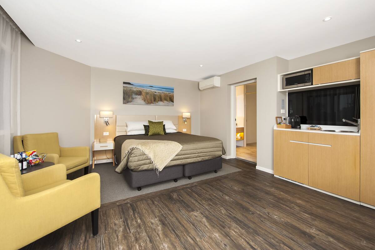 A modern hotel room with a large bed, a sofa, a desk, and a kitchenette.