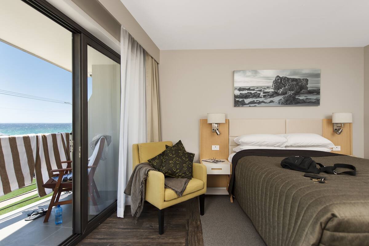A hotel room with a balcony, a bed, a chair, a nightstand, and a painting.