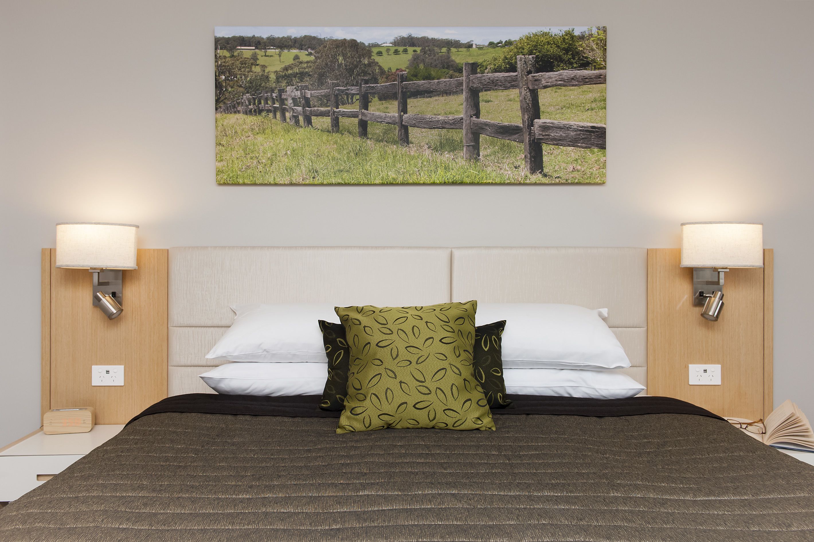 A neatly made bed with a patterned pillow, two bedside lamps, and a framed picture of a fence on the wall.