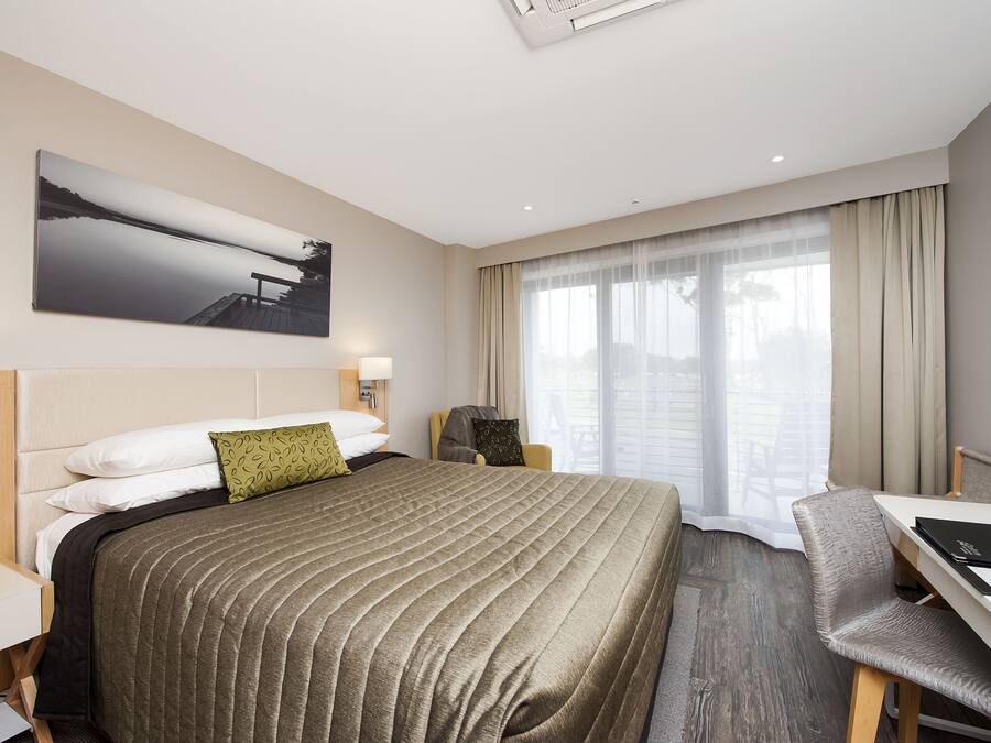 Quarto de hotel com uma cama grande, uma escrivaninha e uma cadeira. Há uma janela com cortinas e um quadro na parede.
