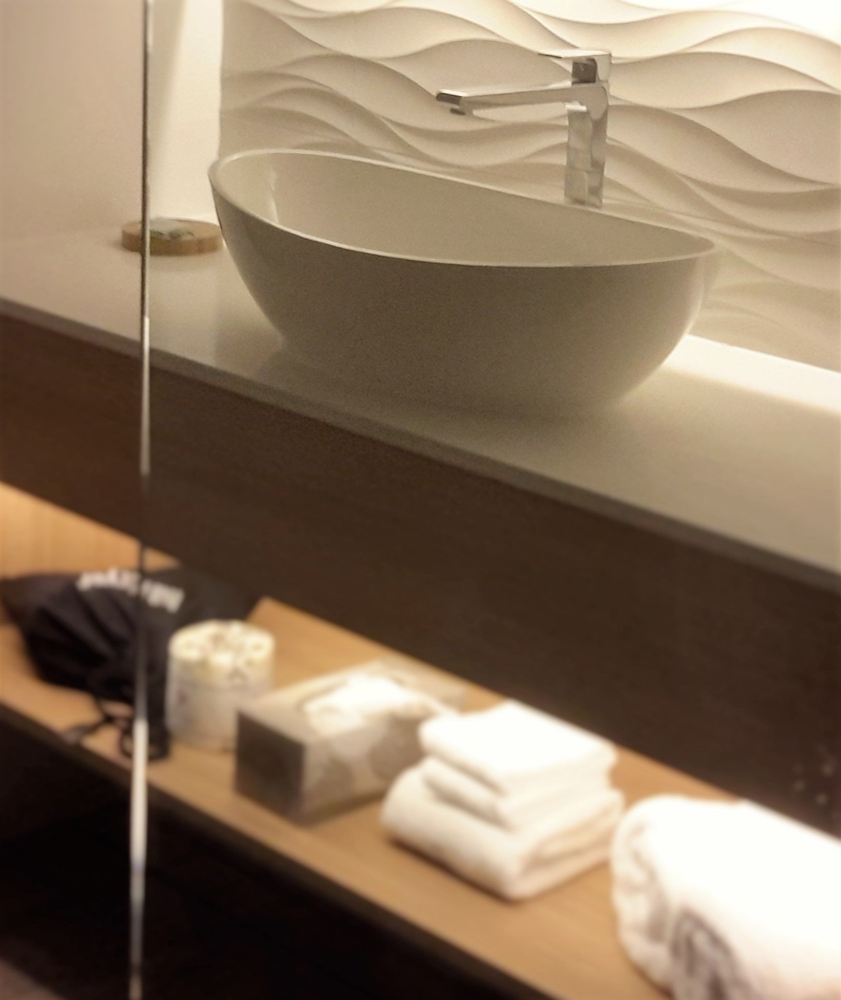 A bathroom with a large, modern vessel sink mounted on a wall with a textured design. There is a wooden vanity with towels and toiletries on it.