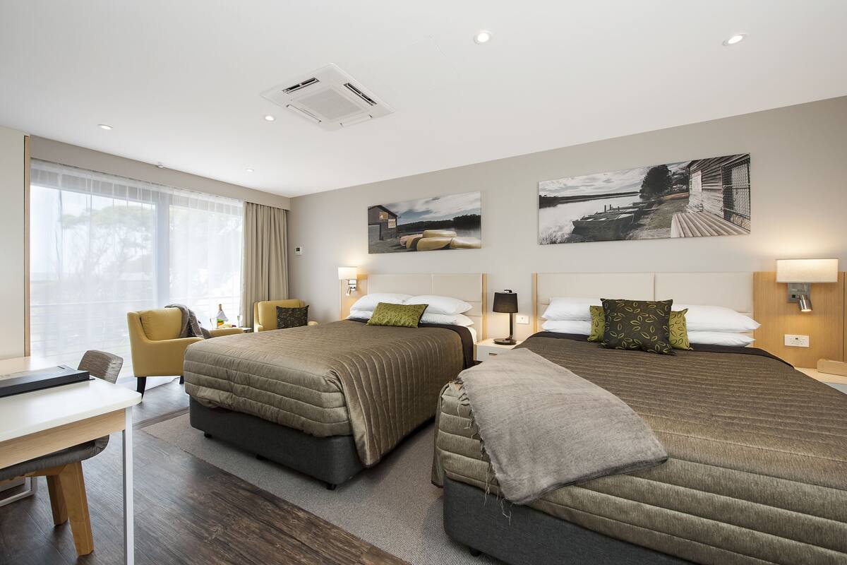 A hotel room with two beds, a desk, and a chair. There are two paintings on the wall and a window with curtains.