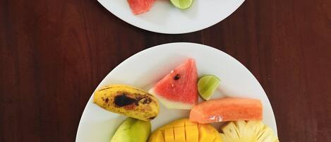Two white plates with sliced fruits including bananas, watermelon, mango, pineapple, and lime on a wooden table.