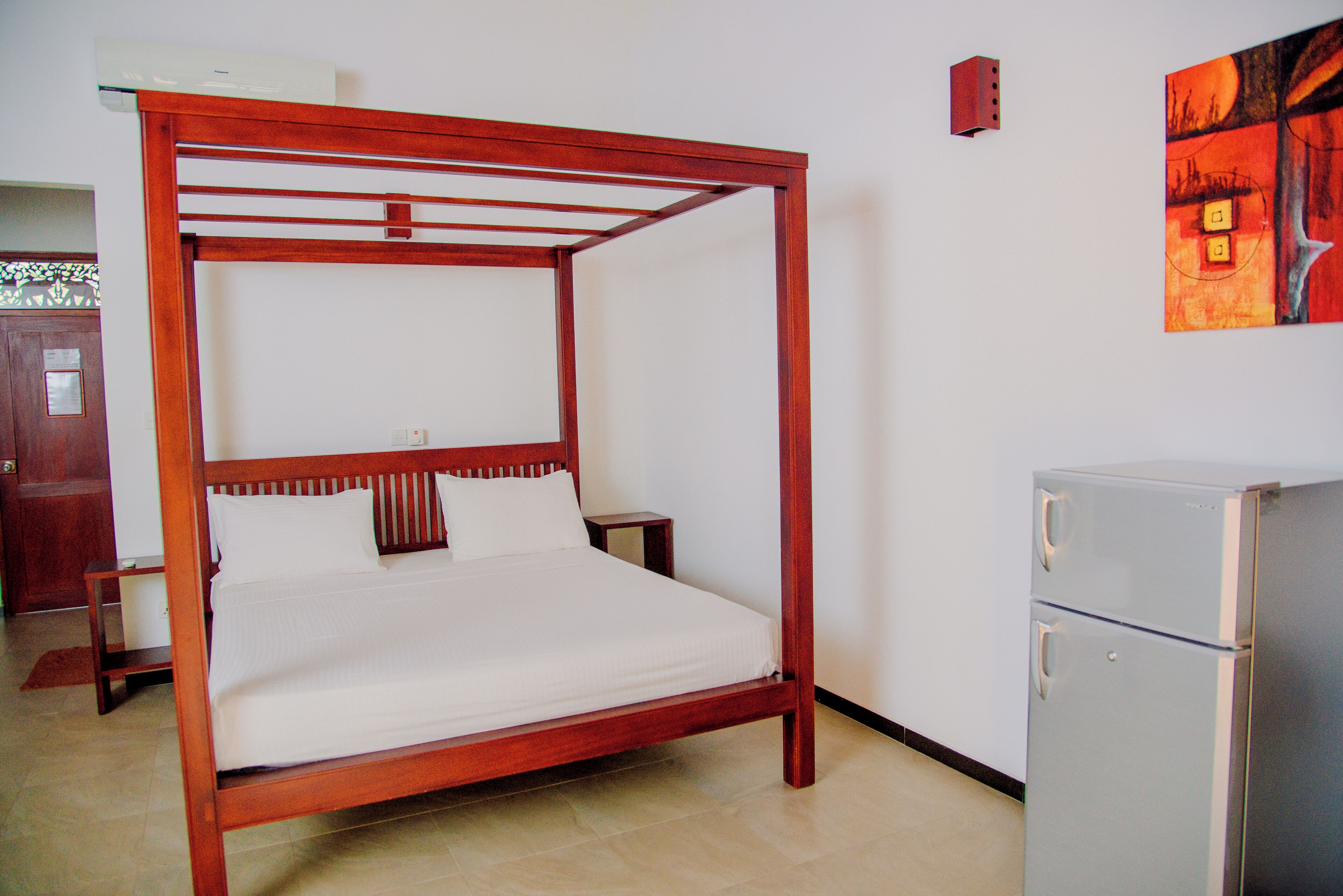 A four-poster bed with a wooden frame and white bedding, a small bedside table, and a refrigerator in a room with a tiled floor and a door in the background.