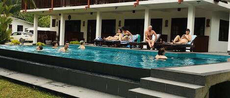 Un área de piscina con un edificio de dos pisos, una terraza cubierta y escalones que bajan a la piscina.