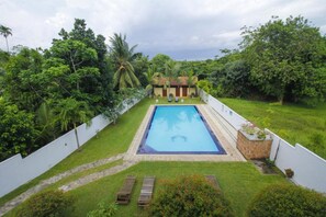Una piscina rectangular rodeada por una cerca blanca, con un edificio pequeño al fondo y abundante vegetación.