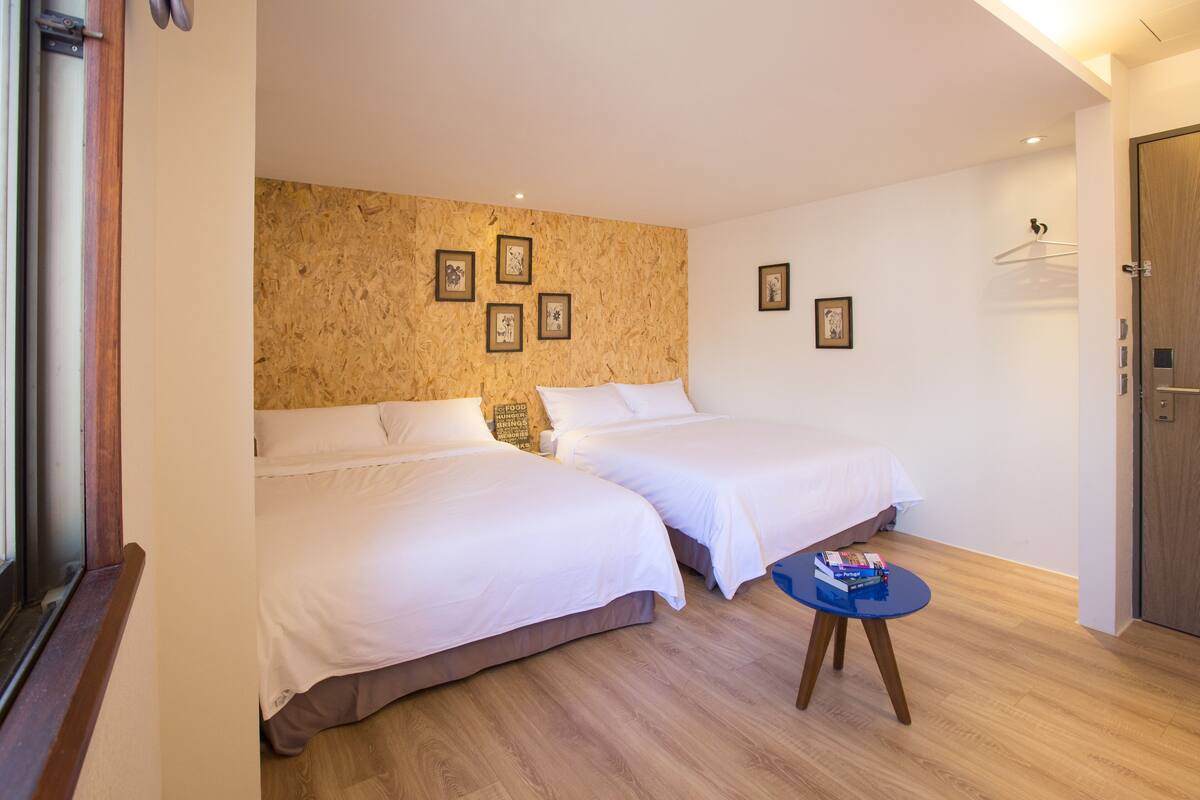 Quarto de hotel com duas camas, uma mesa redonda pequena e uma parede de madeira com quadros.