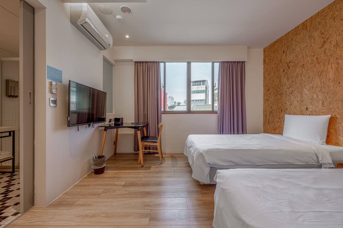 Quarto de hotel com duas camas, uma escrivaninha com cadeira, televisão e uma janela com cortinas.