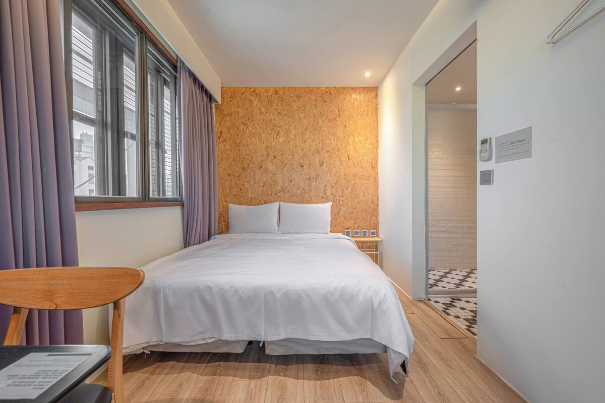Quarto de hotel com cabeceira de madeira, cama com roupa de cama branca, uma cadeira de madeira, uma mesinha e vista para a cidade pela janela.