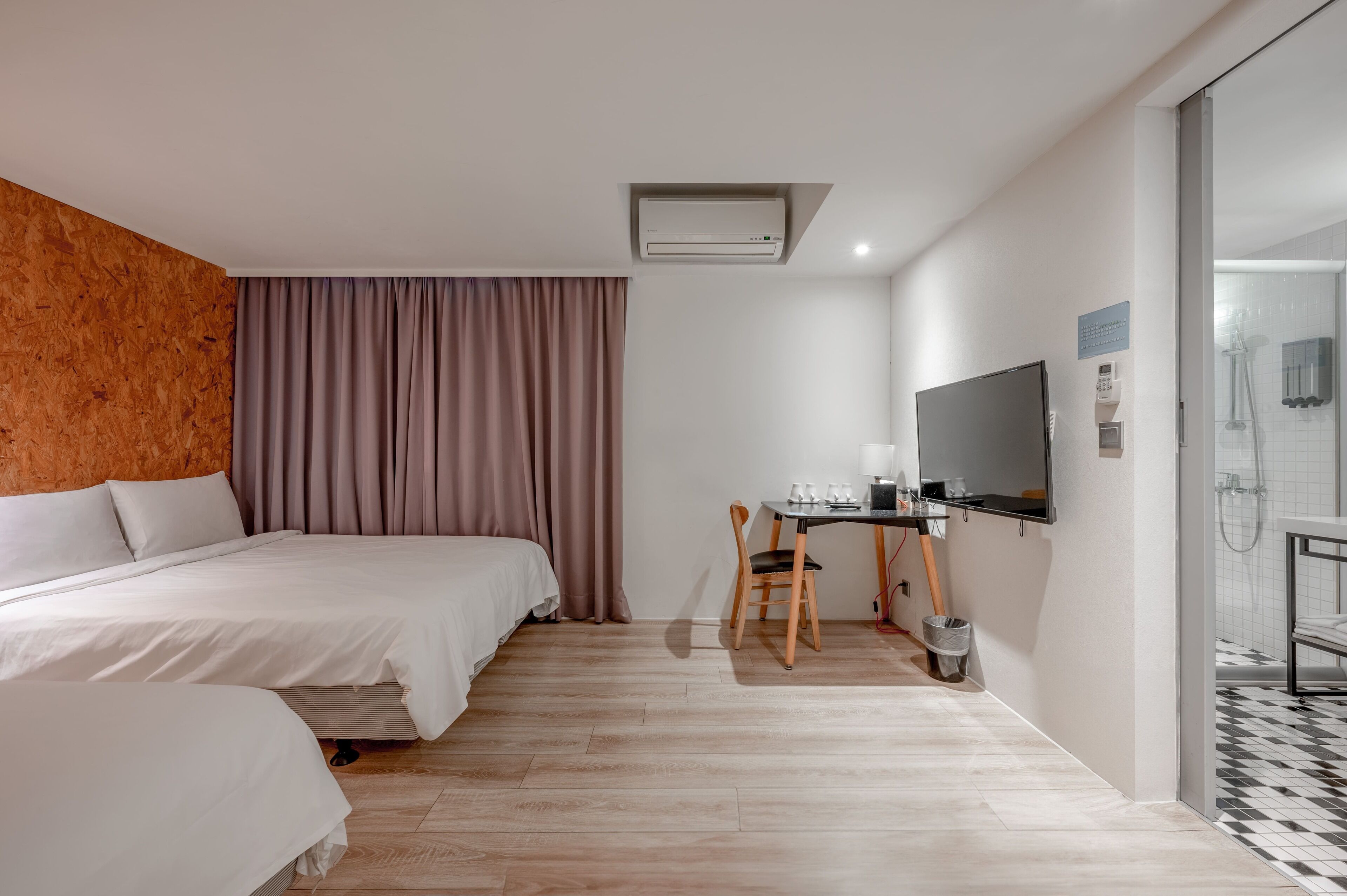 A hotel room with two beds, a desk with a chair, a TV, and a shower area.