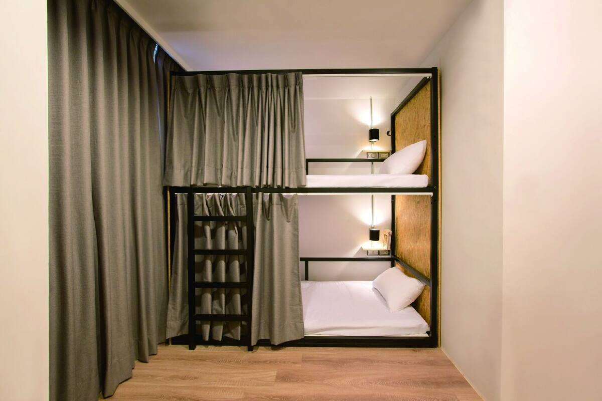 Quarto compacto com cama de solteiro, uma pequena mesa de cabeceira com luminária e cortina.