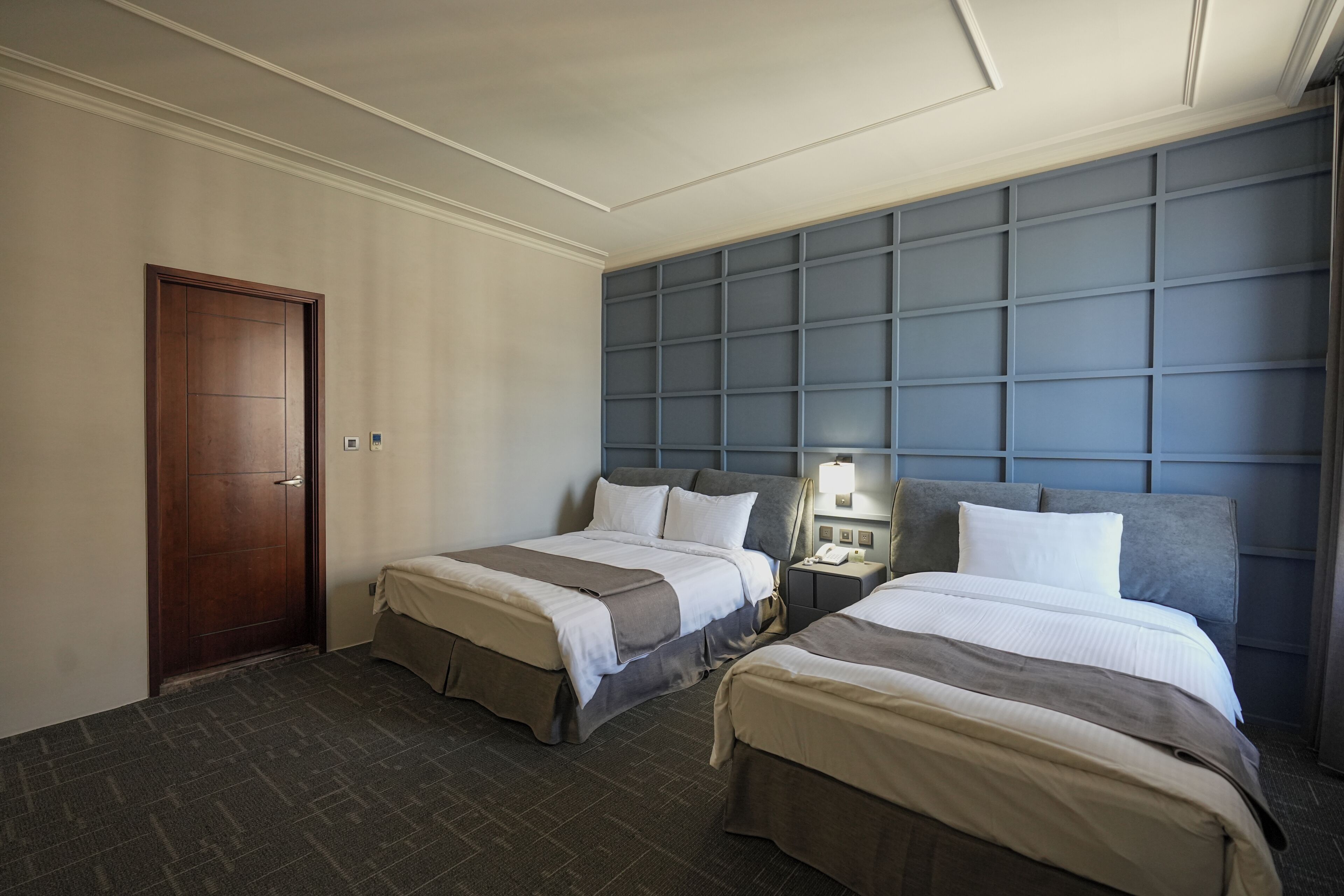 Een hotelkamer met twee bedden, een houten deur, een blauw hoofdeinde en een nachtkastje met een lamp.