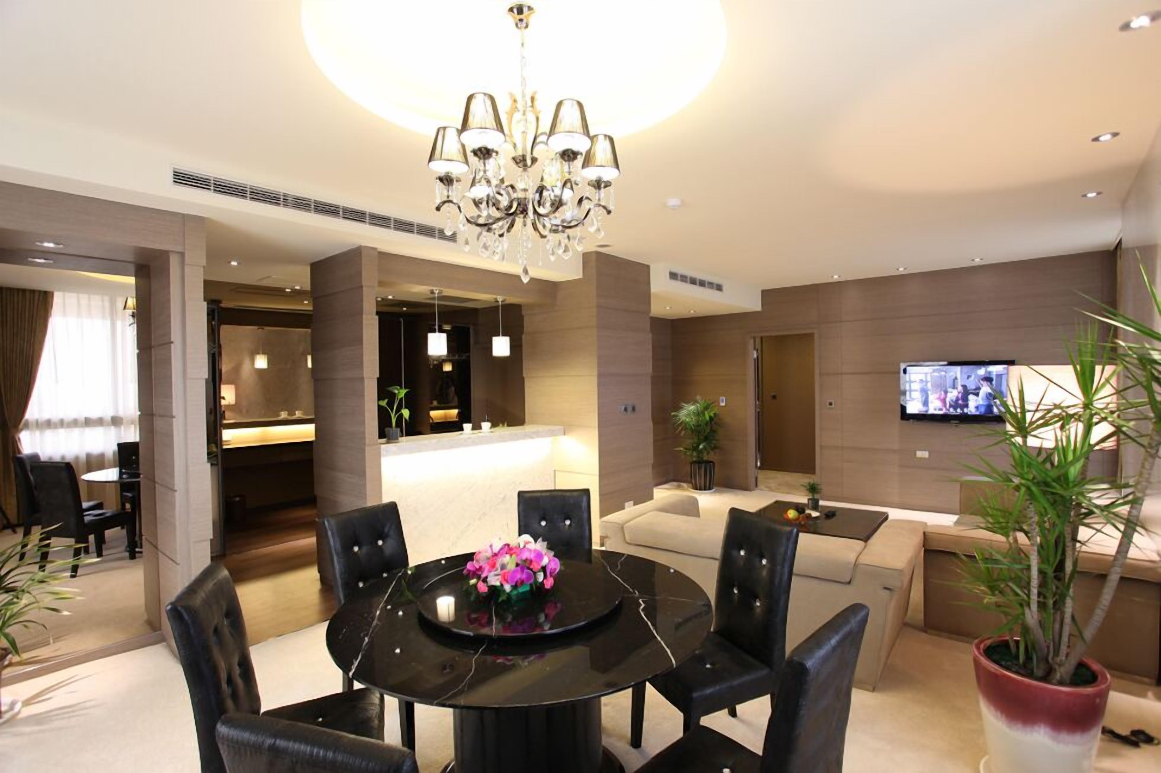 A modern hotel lobby with a round dining table, black chairs, a chandelier, a sofa, a TV, and a potted plant.