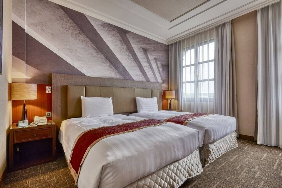 Ein Hotelzimmer mit zwei Betten, einem großen Fenster mit Vorhängen, einem hölzernen Kopfteil und einer strukturierten Decke.