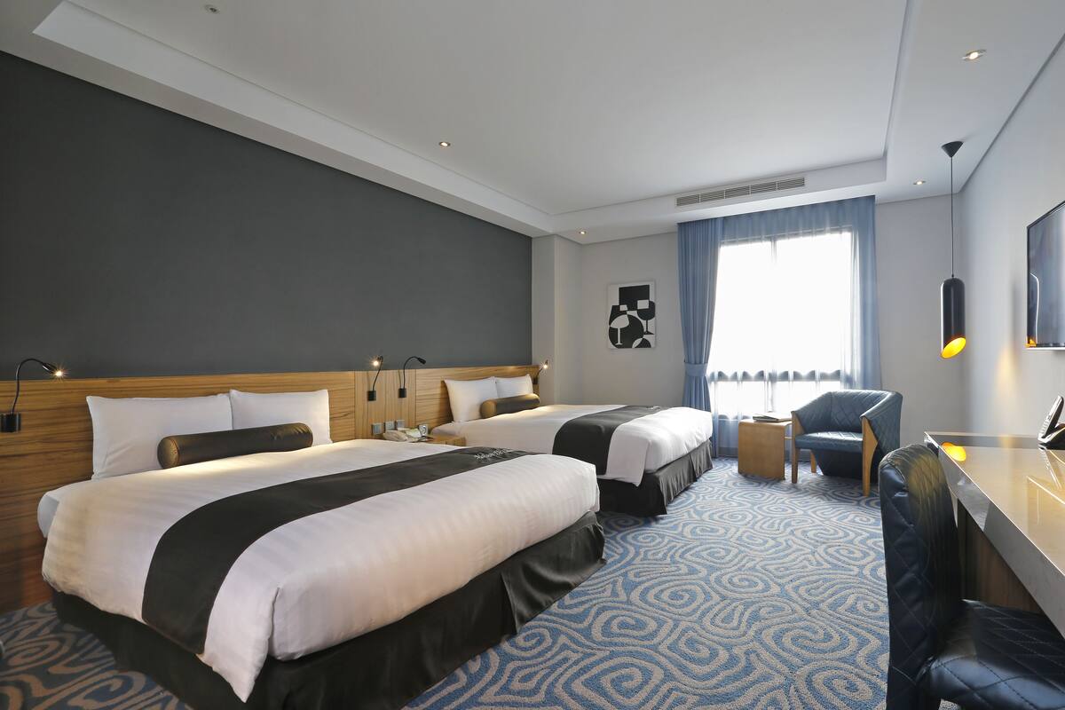 Ein Hotelzimmer mit zwei Betten, einem Schreibtisch, einem Stuhl und einem großen Fenster mit Vorhängen.