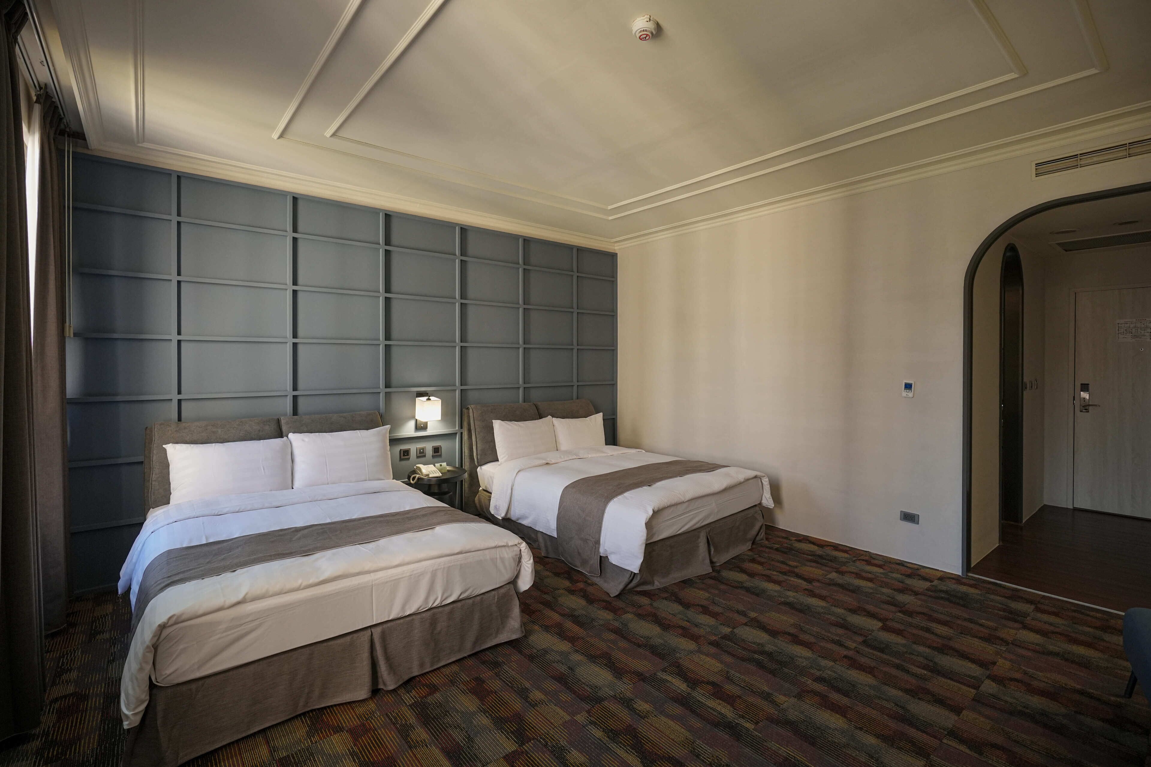A hotel room with two beds, a patterned carpet, a large headboard, and a door to the right.