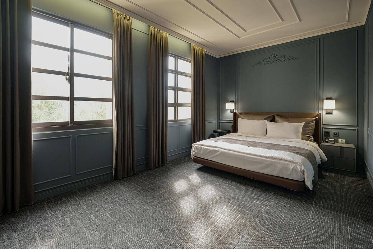Ein Schlafzimmer mit einem großen Bett, zwei Nachttischen, einem Beistelltisch, einem Fenster mit Vorhängen und einer Wandleuchte.