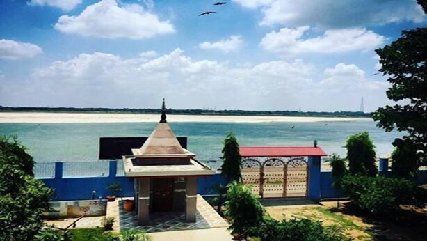 A view of a beach with a small building with a red roof and a white wall in the foreground. - Shri Yoga Mandir Guest House (Varanasi)
