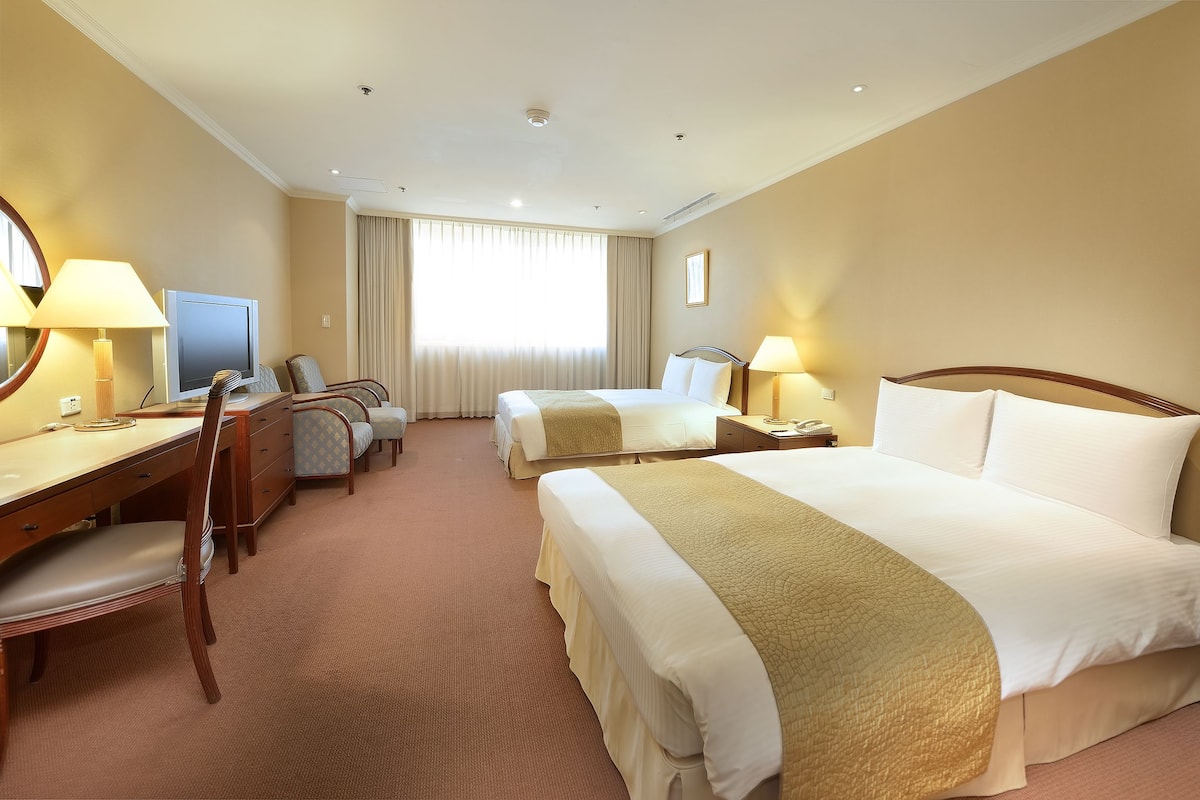 Una habitación de hotel con dos camas, un escritorio con una silla, un televisor y una ventana con cortinas.