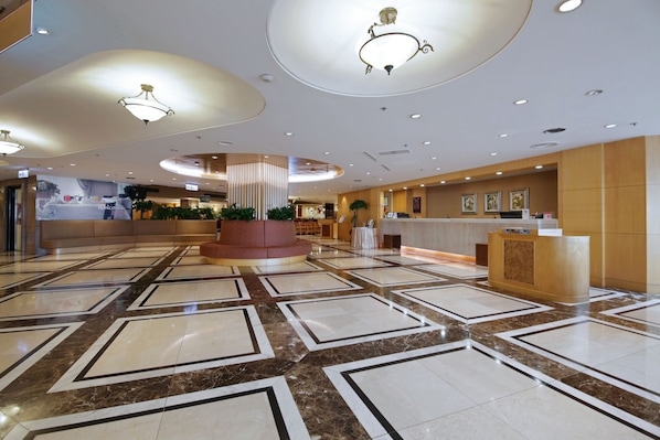 A spacious lobby with a marble floor, a reception desk, and a large central column with plants.