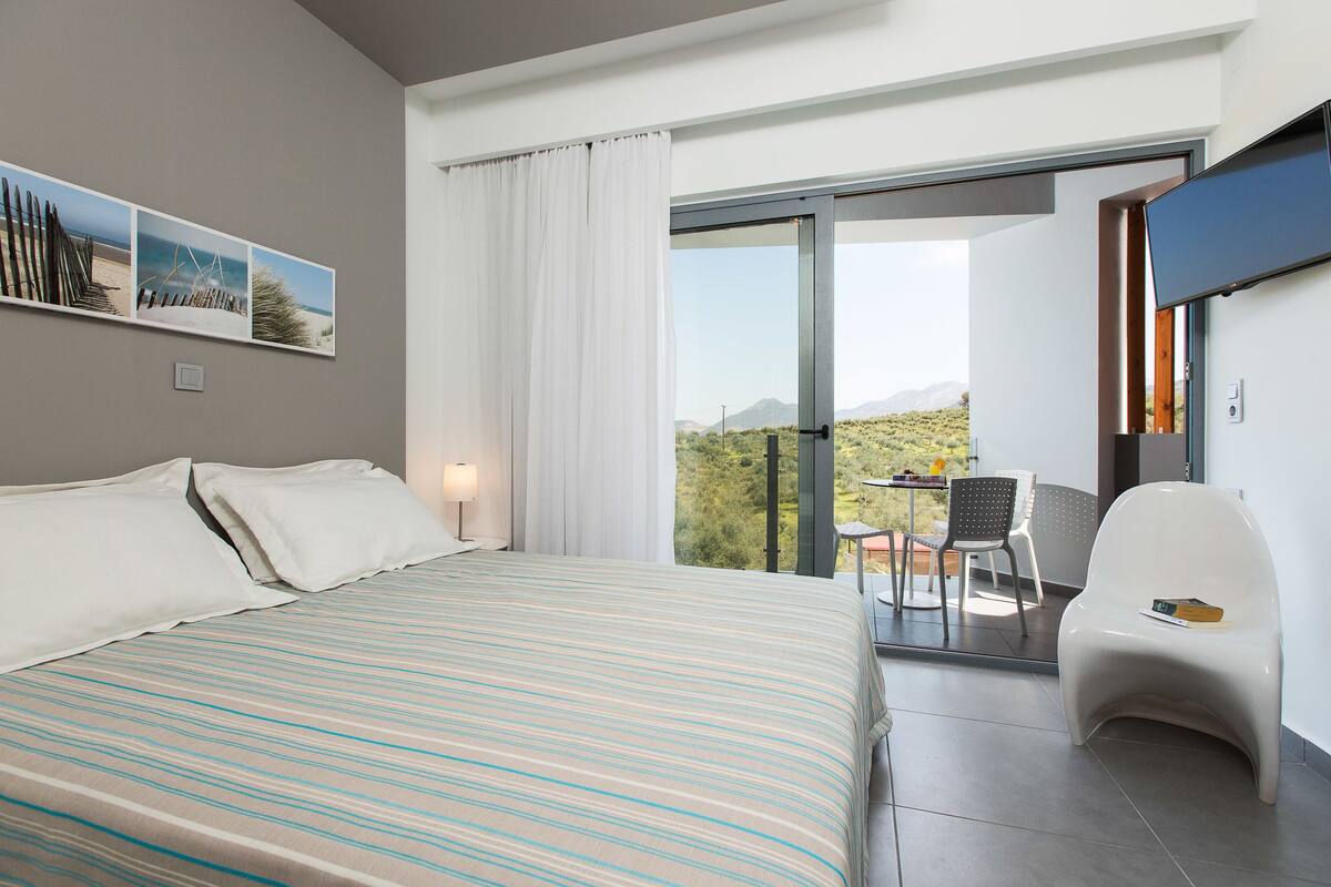 A modern hotel room with a large bed, a balcony with a table and chairs, and a view of the desert landscape.