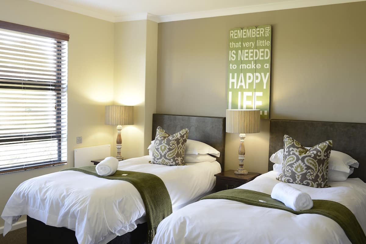 A hotel room with two beds, each with white linens and green throw blankets. There are two bedside lamps, a wall-mounted clock, and a motivational quote on the wall.