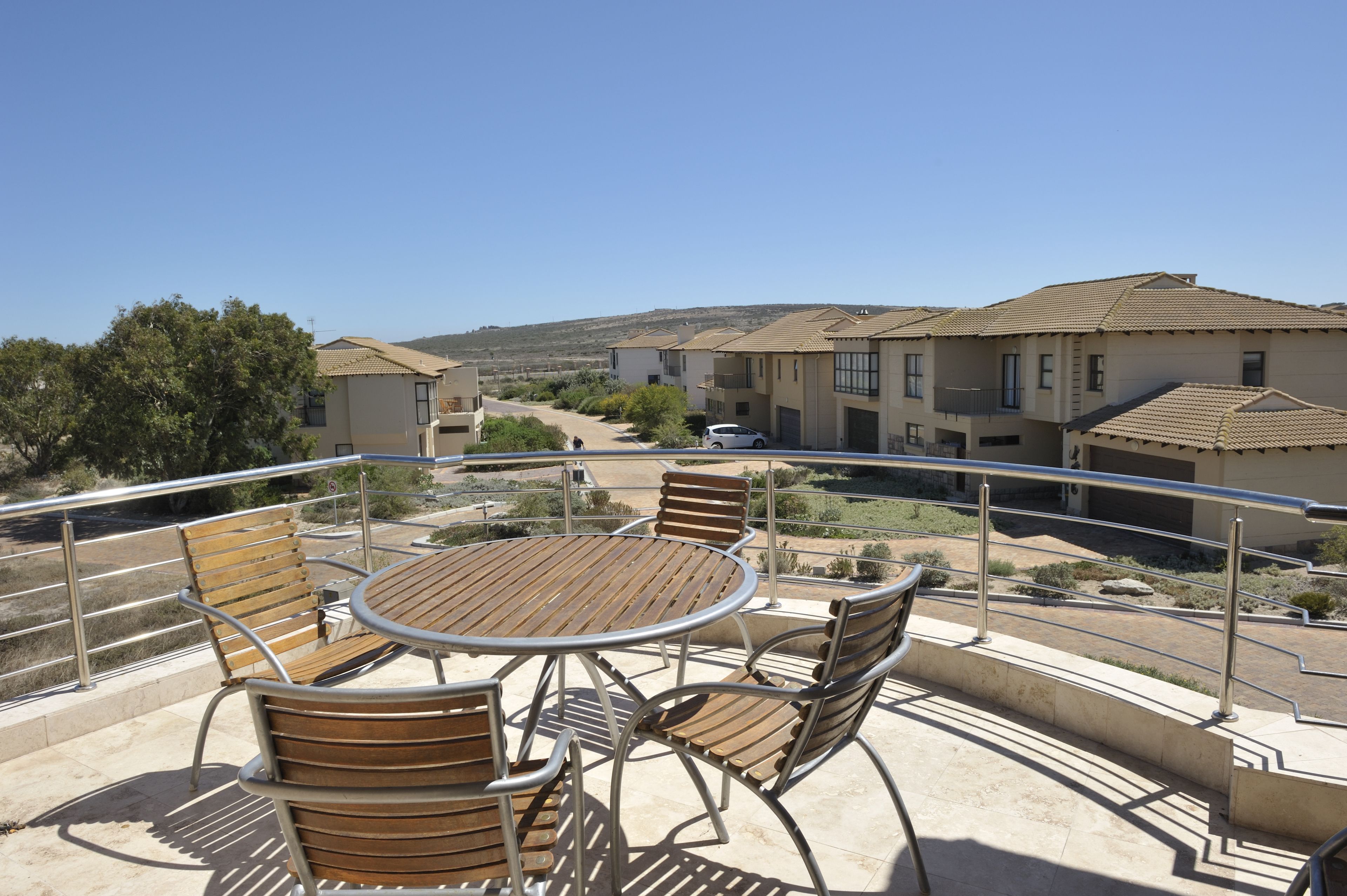Un balcon avec une table ronde et des chaises, donnant sur un quartier résidentiel avec des maisons et un ciel dégagé.