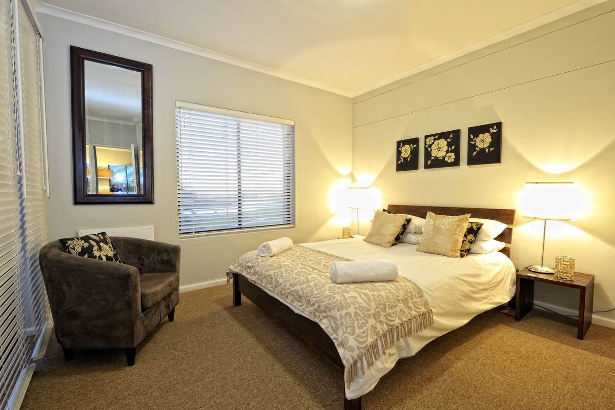 A bedroom with a large bed, two bedside lamps, a chair, a mirror, and three wall-mounted floral decorations.