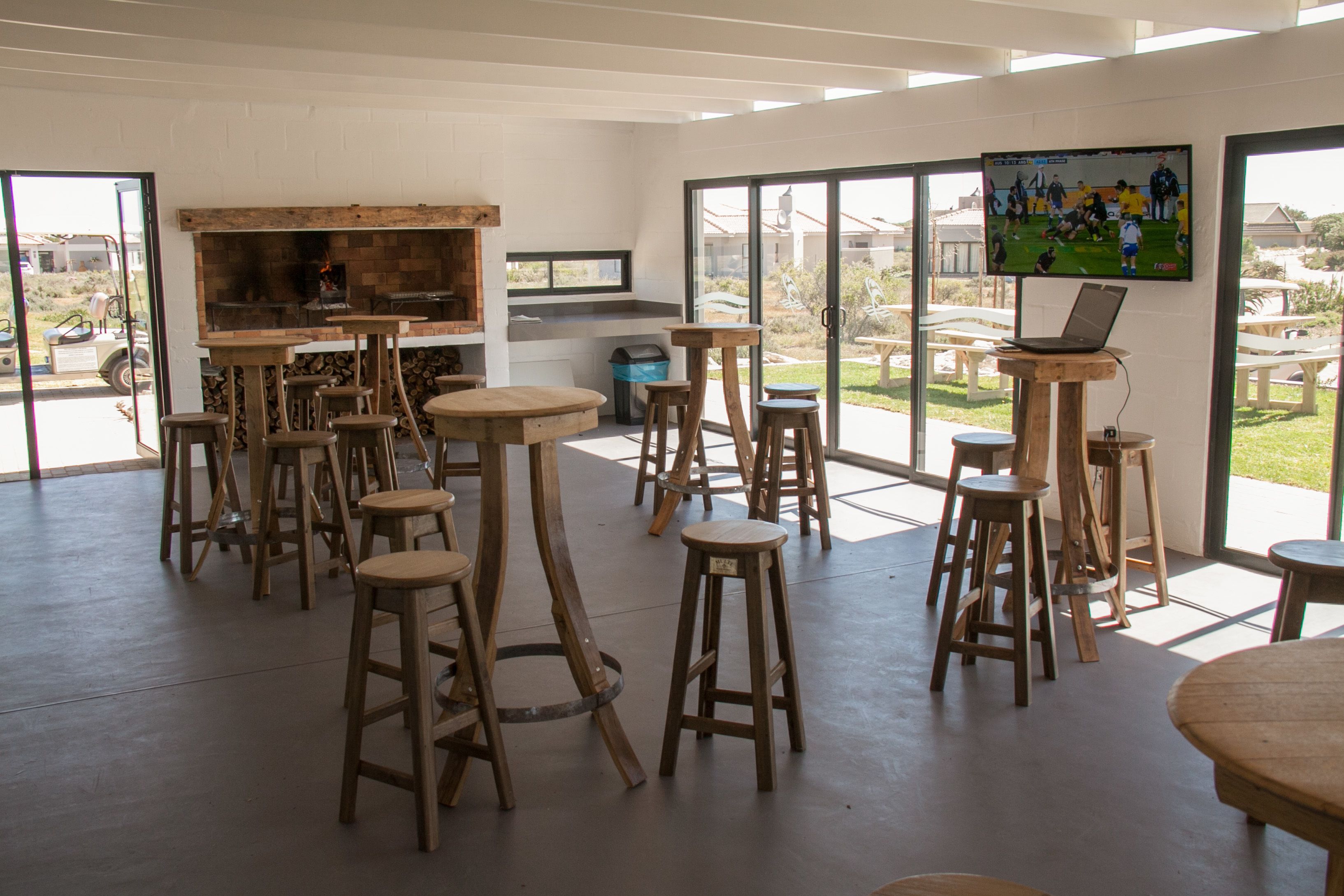A spacious indoor area with multiple wooden stools and tables, a large TV screen, and a laptop on a wooden stand.