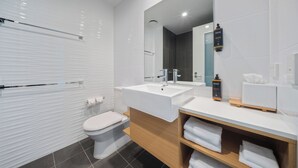 A modern bathroom with a white toilet, a large sink, and a mirror. There are towels on a shelf and a soap dispenser on the sink.