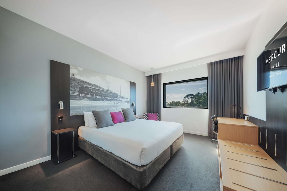 Una habitación de hotel moderna con una cama grande, un escritorio, un televisor y una ventana con vistas a árboles y un edificio.