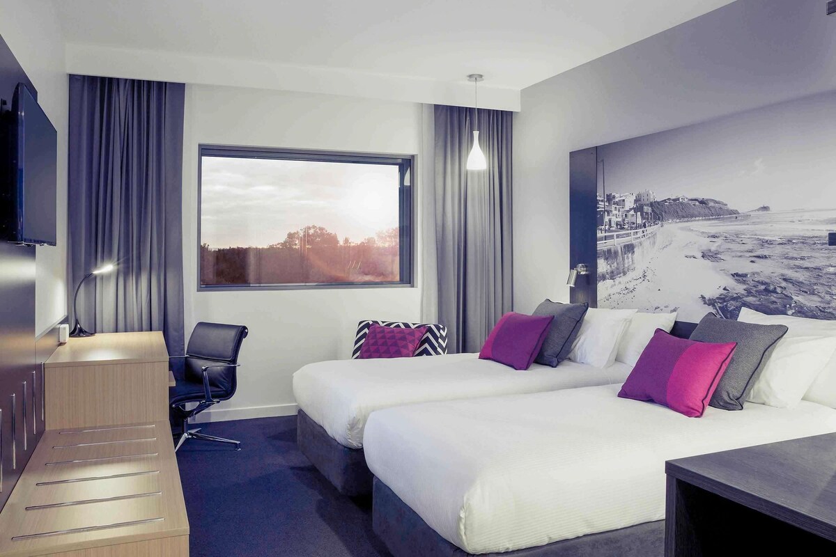A modern hotel room with two beds, a desk, a chair, a TV, and a large window with a beach view.