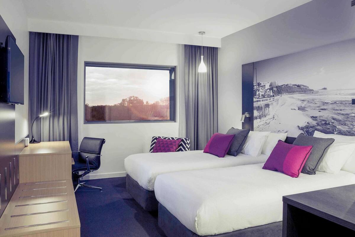 Una habitación de hotel moderna con dos camas, un escritorio, una silla, un televisor y un gran ventanal con vista al mar.